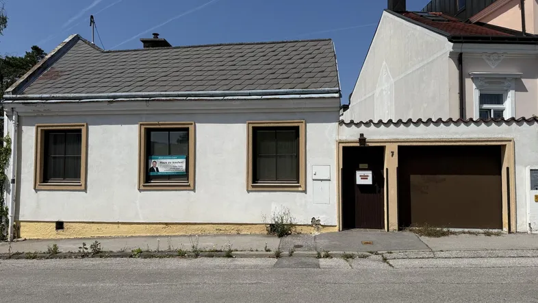 Einfaches Haus mit weißer Fassade, grauen Dachziegeln und integrierter Garage an einer Straße.