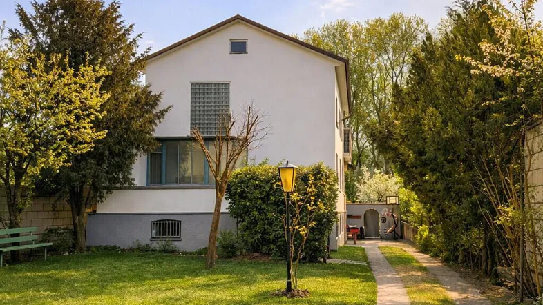 Weißes Einfamilienhaus mit großem Garten, Zufahrt und umgebender Hecke unter blauem Himmel.