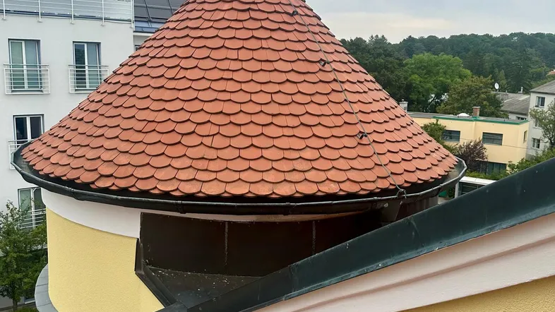 Nahaufnahme des Turmdachs mit Terrakotta-Ziegeln und Wetterhahn, umgeben von weiteren Gebäuden.