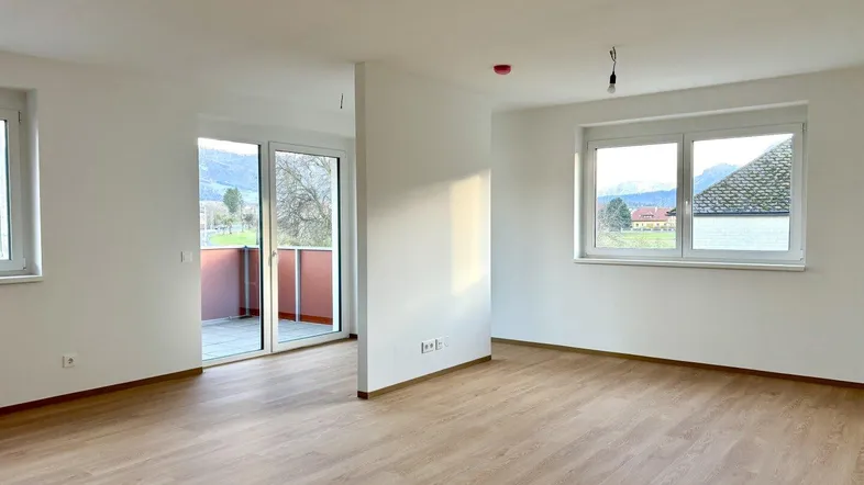 Helles, leeres Wohnzimmer mit Laminatboden und Zugang zu einem Balkon, mit Blick auf die Berge.