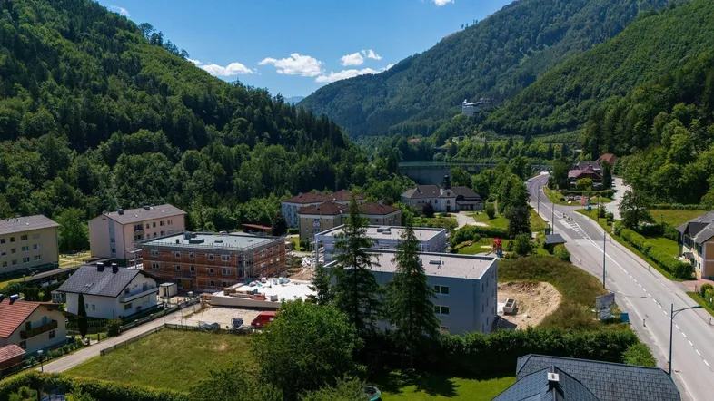 Luftaufnahme eines Tals mit Wohngebäuden, einer Straße und grünen Bergen im Hintergrund.