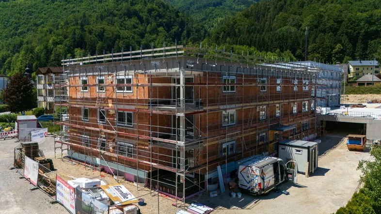Neubau im Rohbauzustand mit Gerüst vor bewaldeter Bergkulisse unter blauem Himmel.