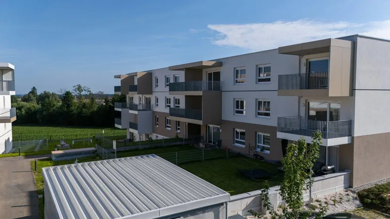 Moderne Wohnanlage mit Balkonen, Gärten und Blick auf grüne Felder unter blauem Himmel.