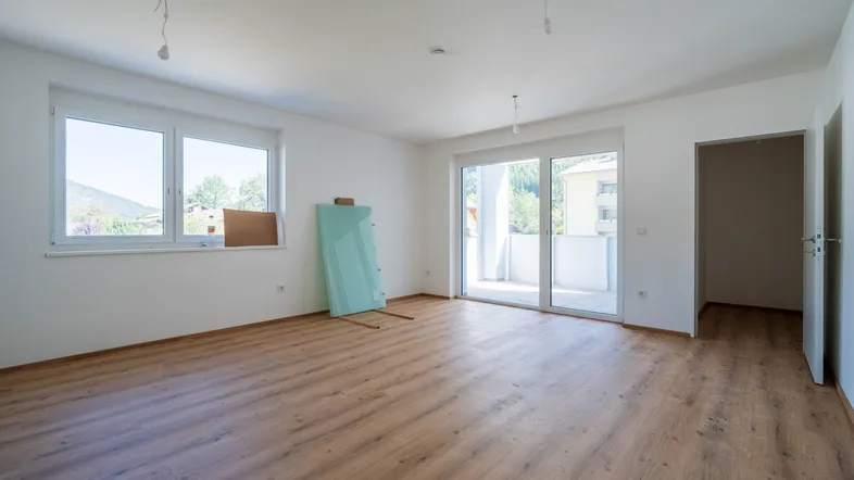 Helles, leeres Zimmer mit Laminatboden, großen Fenstern und Zugang zu einem Balkon.