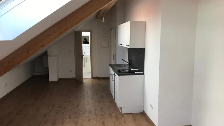 Heller Dachbodenraum mit sichtbaren Holzbalken, großem Dachfenster und integrierter Küchenzeile.
