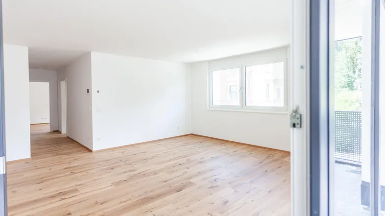 Heller, leerer Wohnraum mit Holzboden und Zugang zu einem Balkon.