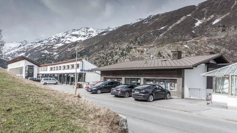 Gebäudeansicht mit geparkten Autos vor einer beeindruckenden Bergkulisse unter bewölktem Himmel.