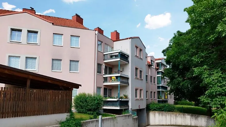 Mehrfamilienhaus mit mehreren Balkonen, Carport und Zufahrt zur Tiefgarage unter blauem Himmel.