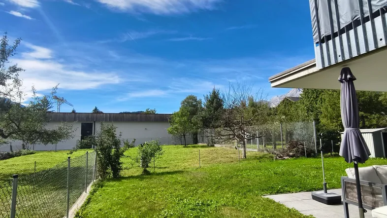 Großer Gartenbereich mit Rasenfläche, Bäumen und einer Terrasse mit Sitzgelegenheiten unter blauem Himmel.