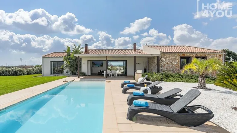 Moderne Villa mit großem Poolbereich, Liegestühlen und mediterraner Bepflanzung unter blauem Himmel.