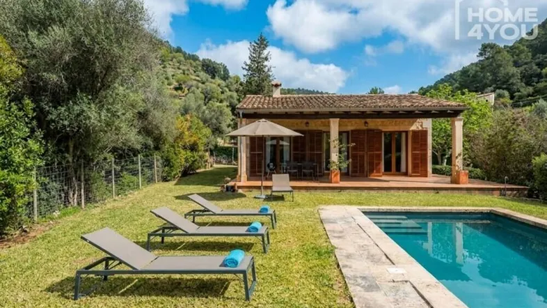 Haus mit Pool und Garten, umgeben von grüner Landschaft unter blauem Himmel.