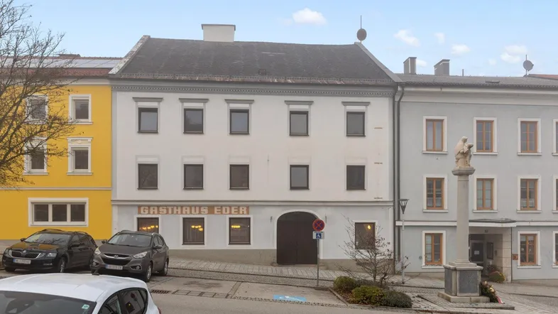 Vorderansicht eines historischen Gebäudes mit Gasthaus im Erdgeschoss und Parkplätzen davor.