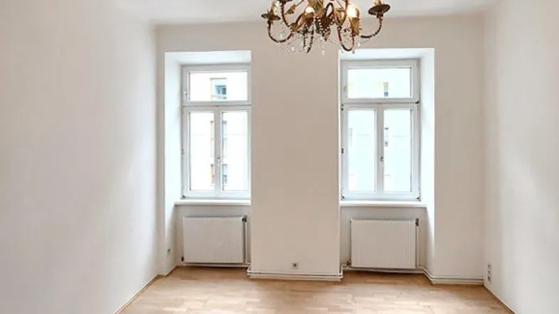 Heller, leerer Raum mit Parkettboden, zwei Fenstern und einem klassischen Kronleuchter an der Decke.