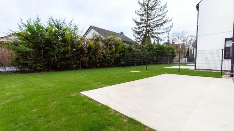 Großer, gepflegter Garten mit Rasenfläche und einer hellen gepflasterten Terrasse, umgeben von Hecken.