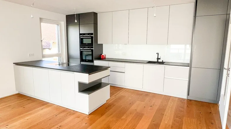 Moderne Einbauküche mit weiß-grauen Fronten, Kochinsel und Holzboden in einem hellen Raum.