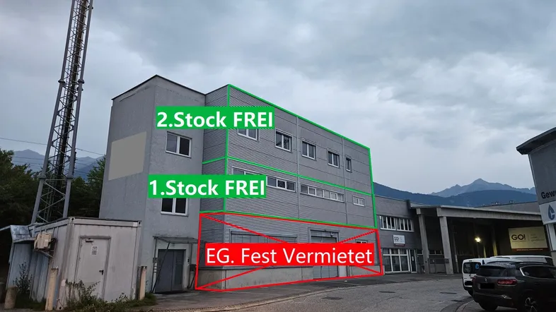 Graues Gewerbegebäude mit Stockwerksangaben und einem Sendemast im Hintergrund unter bewölktem Himmel.