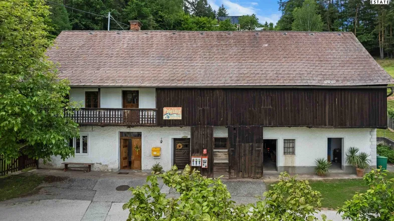 Traditionelles Bauernhaus mit weißer Fassade, Holzbalkon und großem Scheunentor, umgeben von grünen Bäumen.