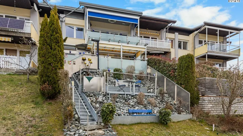 Mehrfamilienhaus mit terrassiertem Gartenbereich, Außentreppe und Balkonen unter blauem Himmel.