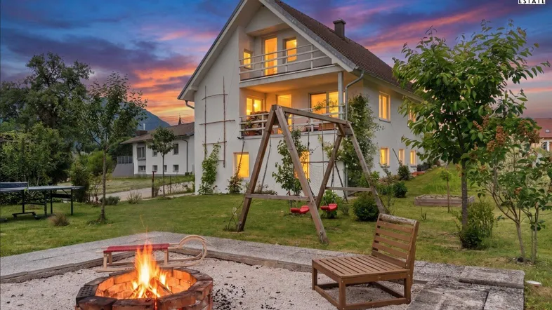 Hausfassade bei Sonnenuntergang mit beleuchteten Fenstern, Feuerstelle und Kinderschaukel im Garten.