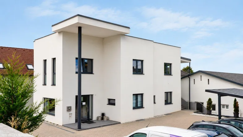 Modernes weißes Gebäude mit Flachdach und dunklen Fensterrahmen, umgeben von einem gepflasterten Parkplatz.