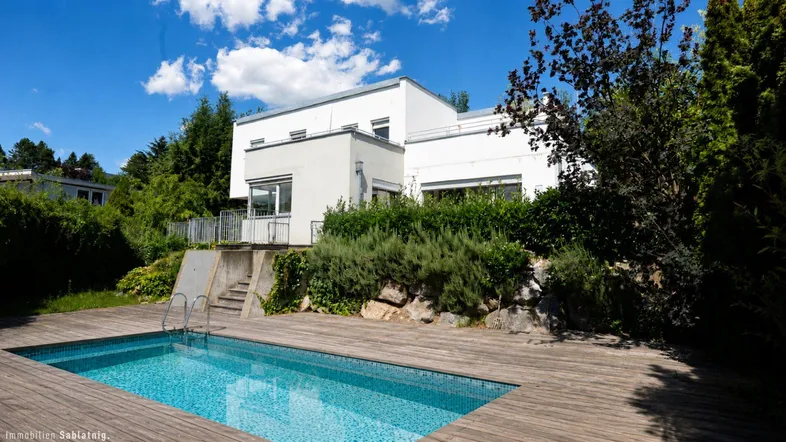 Modernes Haus mit Swimmingpool und Holzdeck, umgeben von viel Grün unter blauem Himmel.