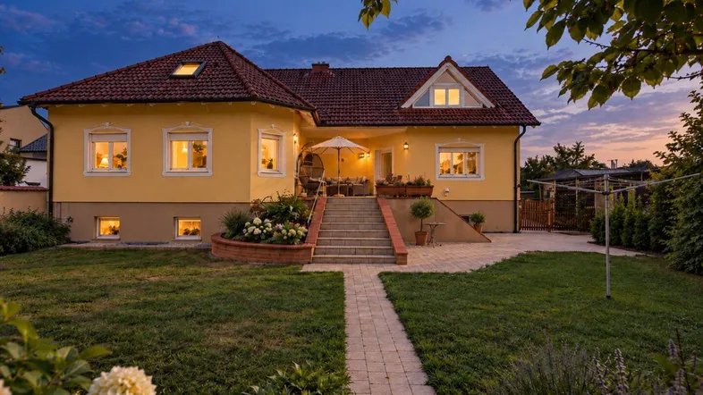 Charmantes gelbes Haus mit rotem Ziegeldach, gepflegtem Garten und einladender Terrasse in der Abenddämmerung.