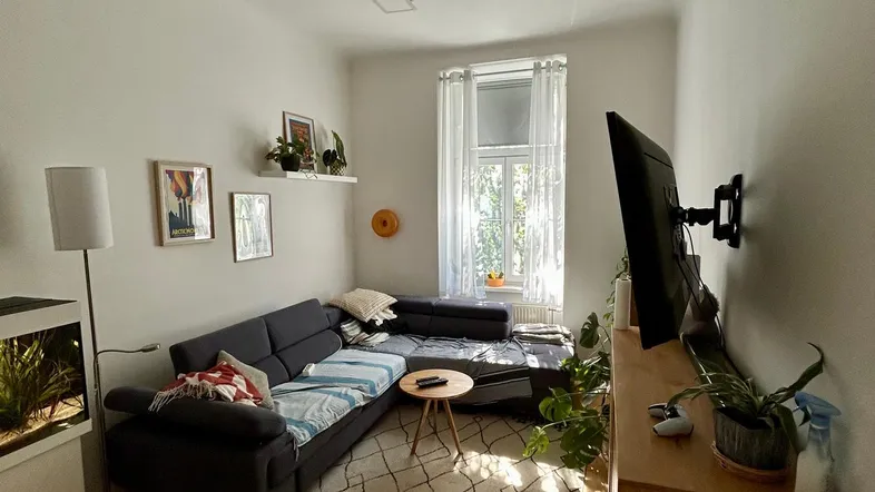 Helles Wohnzimmer mit gemütlichem Ecksofa, Holzboden und einem Fenster, das viel Tageslicht hereinlässt.