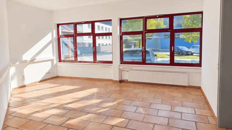 Heller, leerer Raum mit Terrakottafliesen und großen Fenstern mit roten Rahmen, ideal für Büronutzung.
