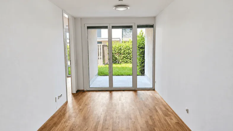 Helles Zimmer mit Holzboden und großen Glasschiebetüren, die zu einer Terrasse und einem kleinen Garten führen.