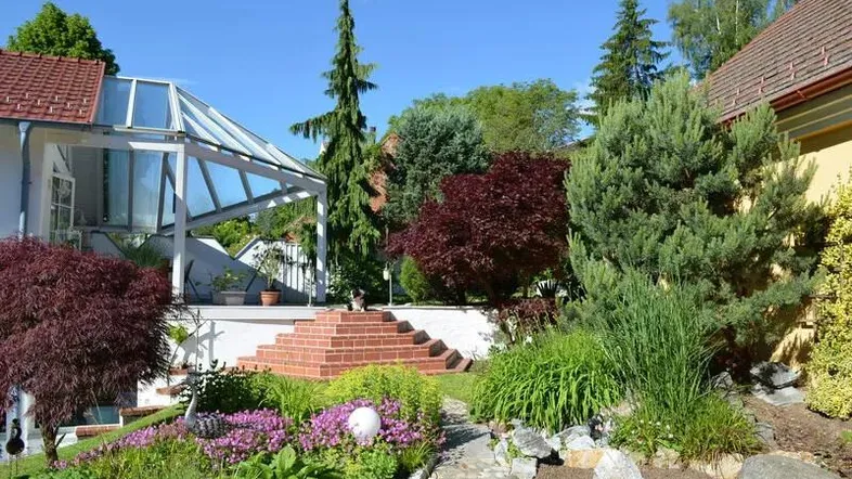 Gepflegter Garten mit vielfältiger Bepflanzung, Steintreppe und einem Wintergarten am Haus.
