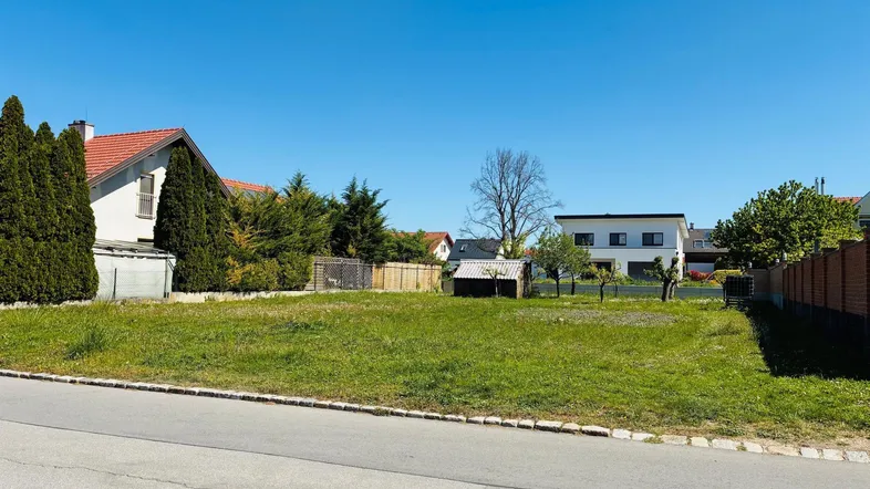 Freies Baugrundstück mit Rasenfläche und umliegenden Wohnhäusern unter klarem blauem Himmel.