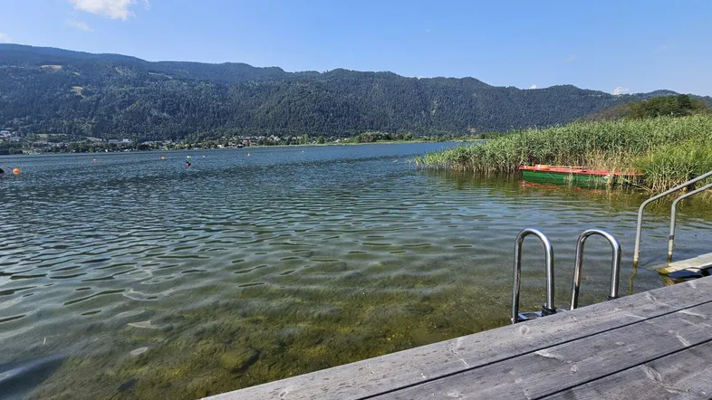Klarer See mit Holzsteg und Bergen im Hintergrund unter blauem Himmel.