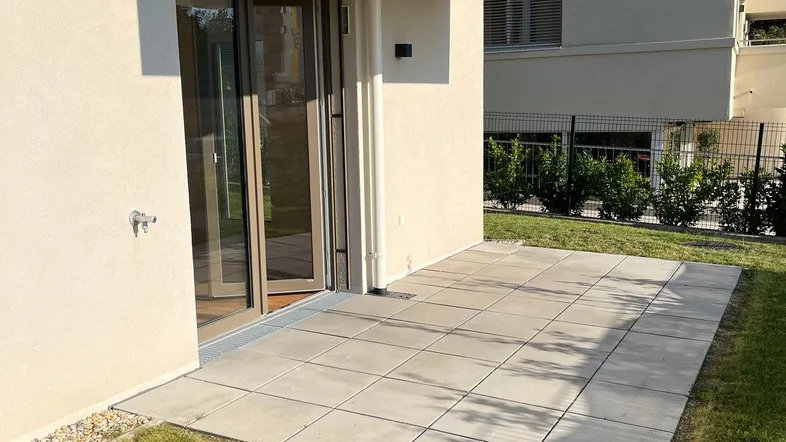 Gepflasterte Terrasse im Erdgeschoss mit direktem Zugang zum Gartenbereich eines modernen Gebäudes.