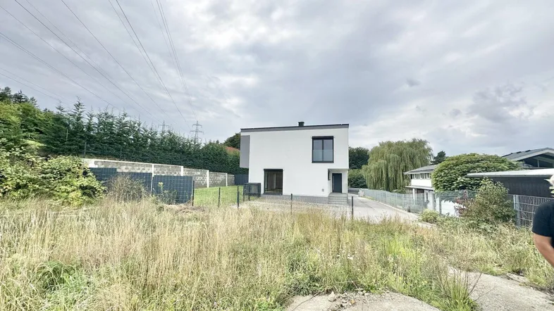 Modernes weißes Einfamilienhaus auf einem großen, noch unbebauten Grundstück unter bewölktem Himmel.