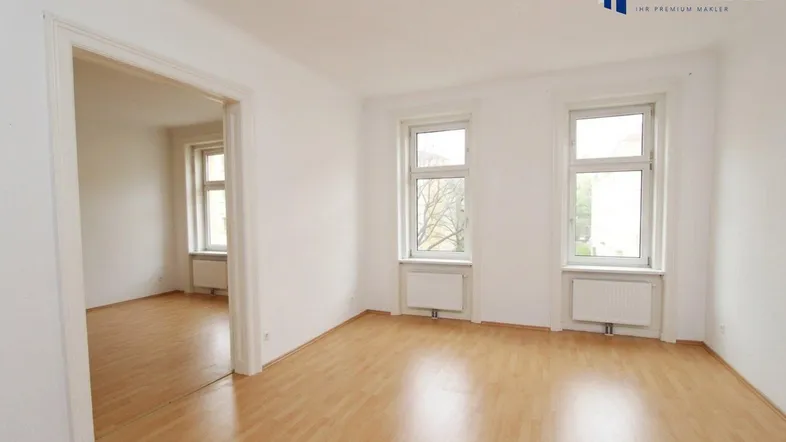 Helles, leeres Zimmer mit Laminatboden und zwei großen Fenstern, das viel Tageslicht hereinlässt.