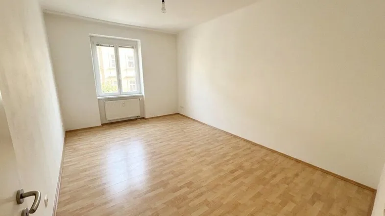 Helles, leeres Zimmer mit Parkettboden und einem Fenster, das Tageslicht hereinlässt.