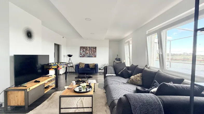 Geräumiges Wohnzimmer mit großem Sofa, Fernseher und weitem Blick durch die Fenster.