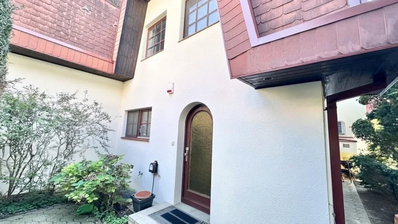 Eingangsbereich eines Hauses mit weißer Fassade, braunem Dach und gepflastertem Vorplatz.