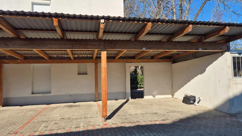 Überdachter Stellplatz mit Holzkonstruktion und gepflastertem Boden vor einem Wohngebäude.