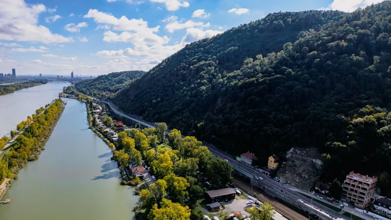 Luftaufnahme der Donau mit bewaldeten Hügeln, Bahnlinie und Stadtpanorama im Hintergrund.