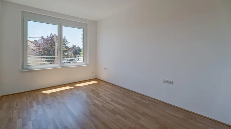 Helles, leeres Zimmer mit Holzparkettboden und großem Fenster, das viel Tageslicht hereinlässt.