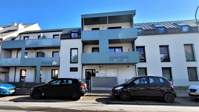 Modernes Mehrfamilienhaus mit weißer Fassade, blauen Balkonen und parkenden Autos auf der Straße.
