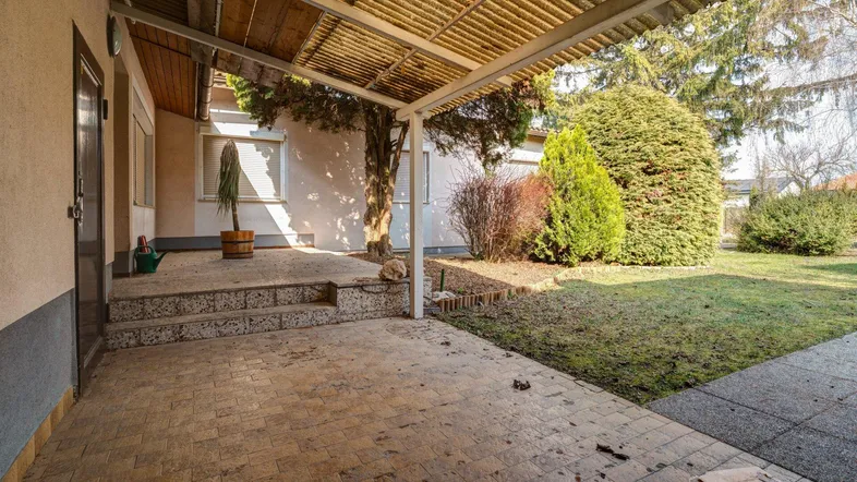 Überdachte Terrasse mit gefliestem Boden und Zugang zum Gartenbereich, umgeben von Grün.