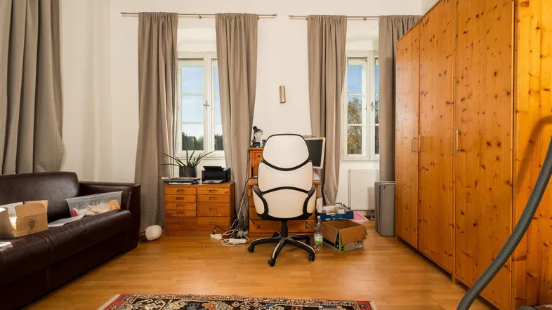 Geräumiges Wohnzimmer mit Holzboden, großem Kleiderschrank und Arbeitsbereich am Fenster.