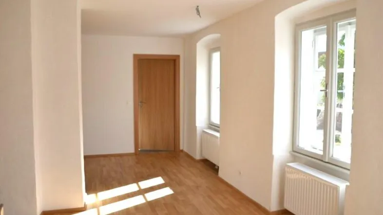 Heller, leerer Raum mit Parkettboden und zwei Fenstern, die viel Tageslicht hereinlassen.