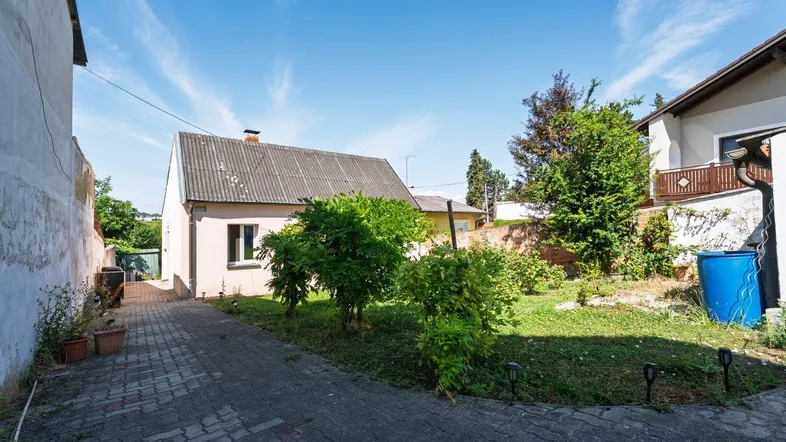 Einfaches Haus mit gepflasterter Zufahrt und kleinem Gartenbereich unter blauem Himmel.