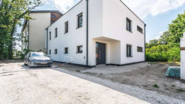 Modernes Einfamilienhaus mit weißer Fassade und dunklen Fenstern, umgeben von Bäumen und einer Kiesauffahrt.