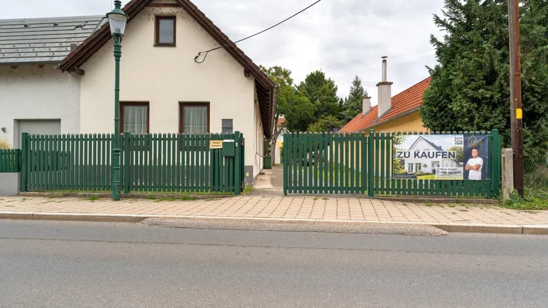Hausfassade von der Straße aus gesehen mit grünem Zaun und einem "Zu kaufen"-Schild.