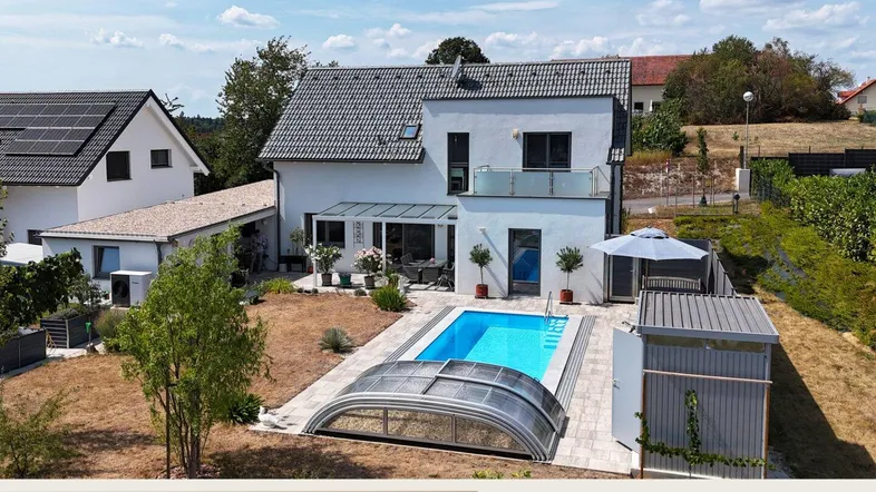 Modernes Haus mit Pool und Gartenbereich, umgeben von ländlicher Landschaft unter blauem Himmel.