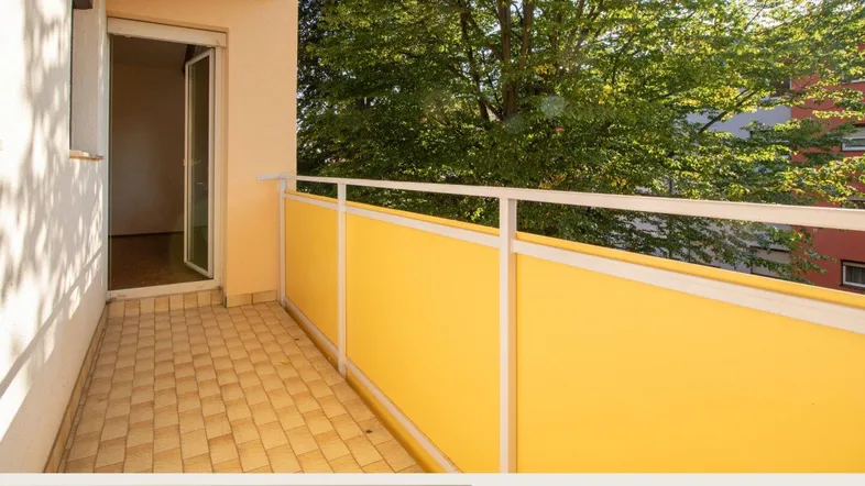 Heller Balkon mit gelbem Geländer und Fliesenboden, umgeben von grünen Bäumen.